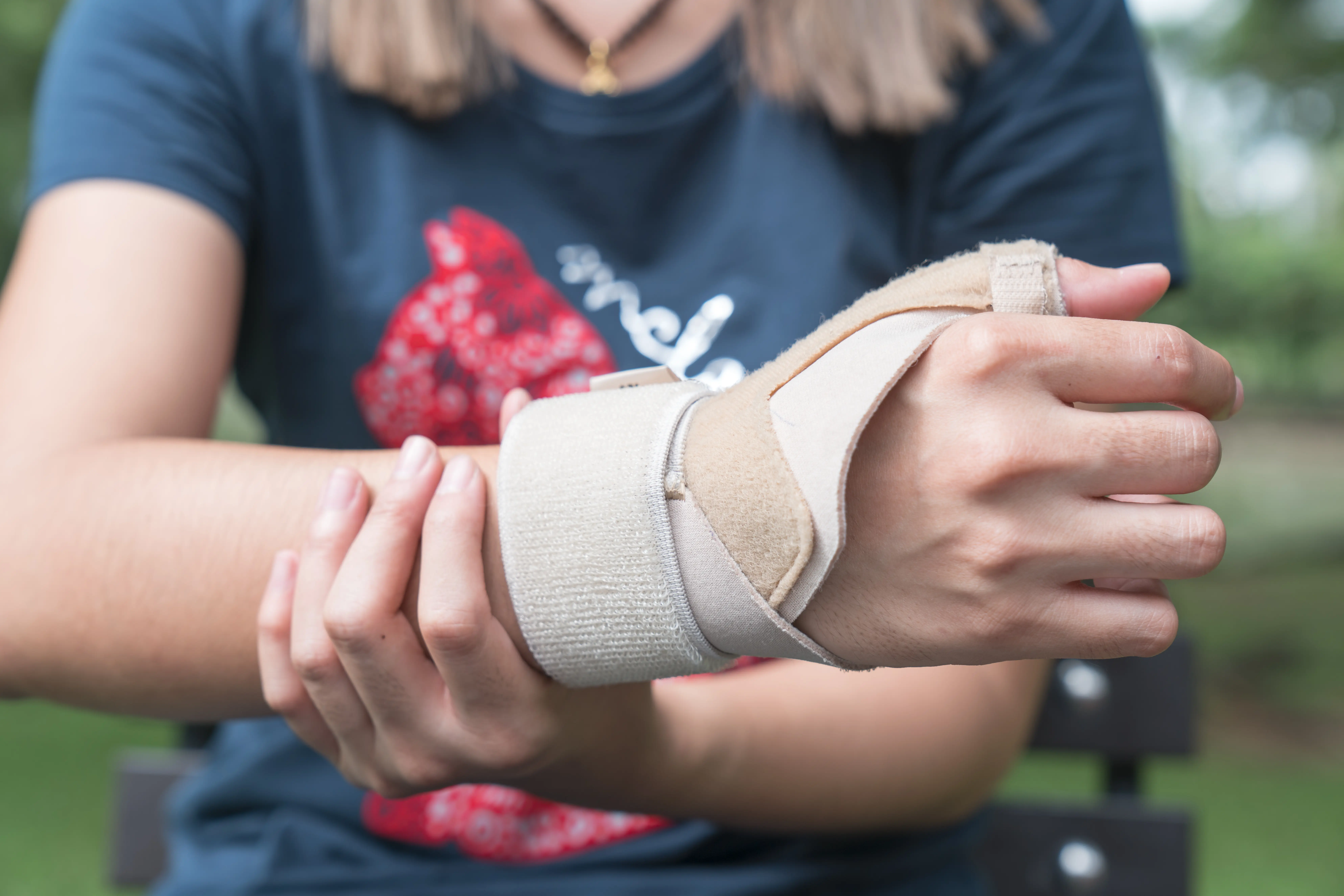 A person wearing a navy shirt shows a bandaged wrist with a beige support brace in a green outdoor setting.
