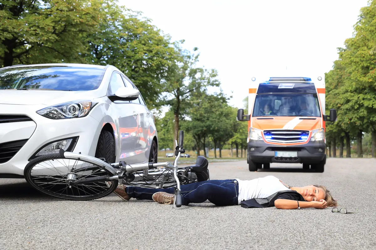 A woman lies on the road next to a fallen bicycle, with a white car nearby and an ambulance approaching.