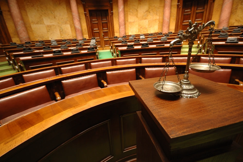Scales of justice on a wooden podium in a grand, empty courtroom with brown leather seating.