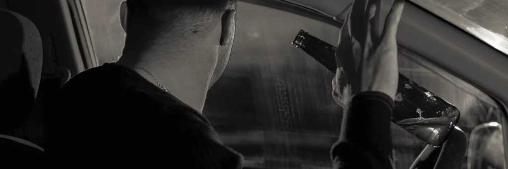 Man holding a beer bottle while sitting in the driver's seat of a parked vehicle at night.