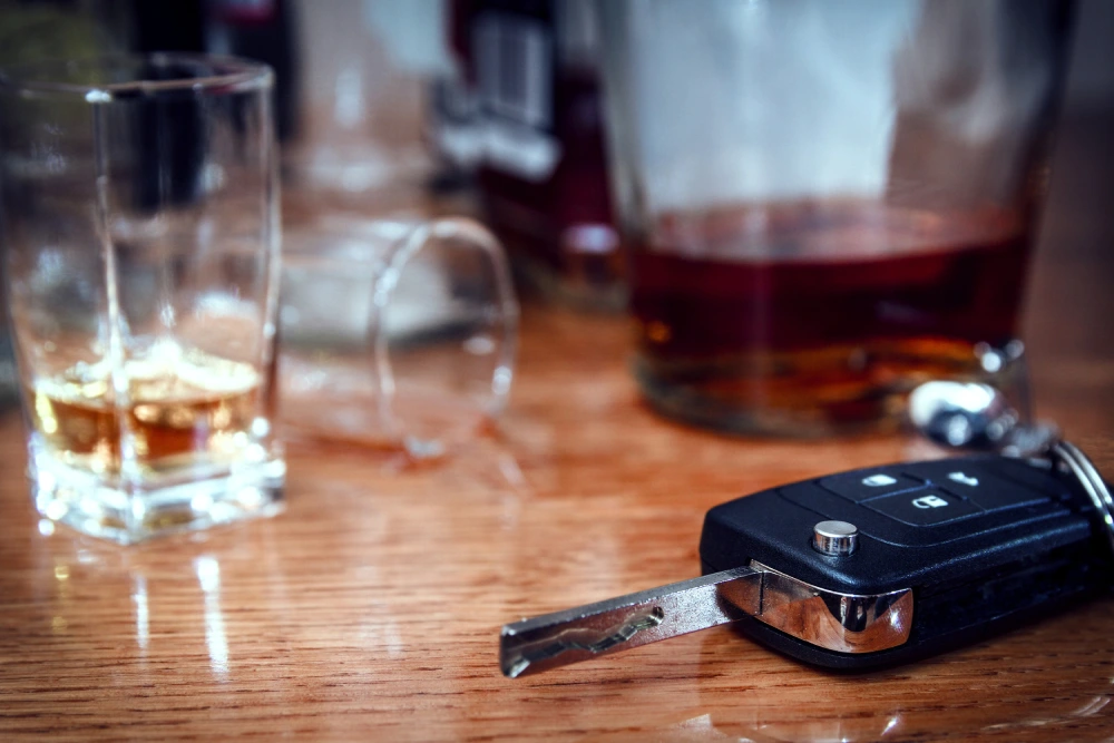 Empty shot glass next to a full glass of whiskey and a car key on a wooden table.