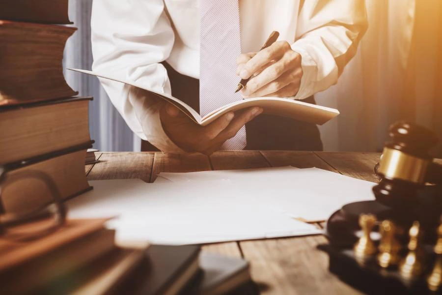 A man in a white shirt and pink tie writes in a notebook, surrounded by law books and a gavel on a wooden table.