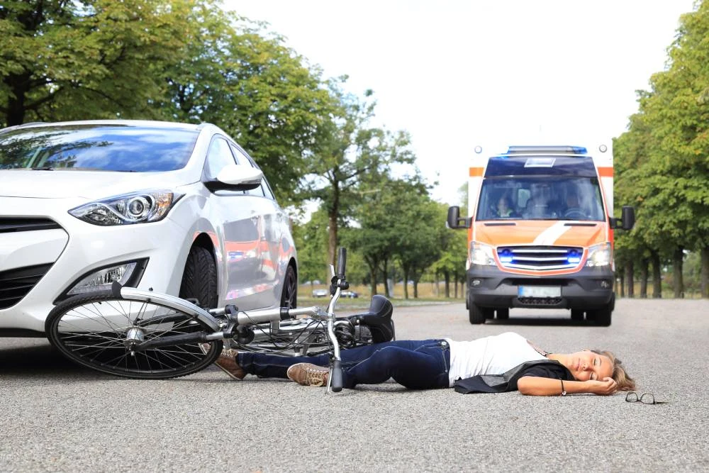 Bicyclist lying on the road near a white car, with an ambulance approaching in the background.