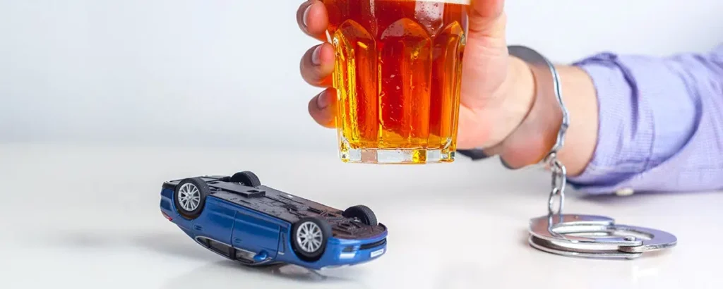 Handcuffed hand holding a glass of alcohol next to an overturned toy car, symbolizing the consequences of speeding and DWI.