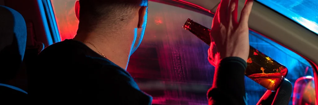 Driver holding a beer bottle inside a car with police lights flashing in the background