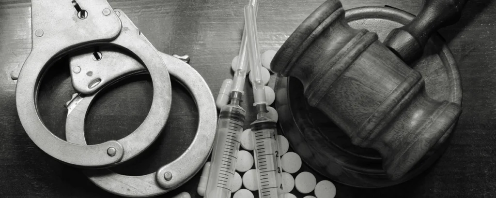 Handcuffs, pills, syringes, and a judge’s gavel arranged on a wooden table, symbolizing drug-related legal consequences.