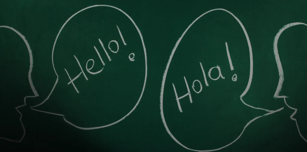 A chalk drawing on a green blackboard features two speech bubbles with the words "Hello!" and "Hola!" written inside them, each accompanied by a silhouette of a person's head in profile. The image represents a bilingual staff, highlighting greetings in English and Spanish.