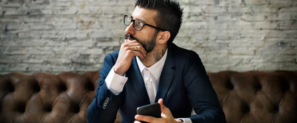 A thoughtful man in a suit holding a smartphone while sitting on a leather couch, symbolizing someone considering options under expungement laws.