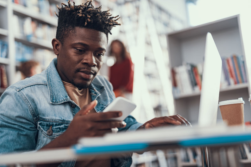 Young Black man in a denim jacket using a smartphone while working on a laptop in a library. Bookshelves in background.