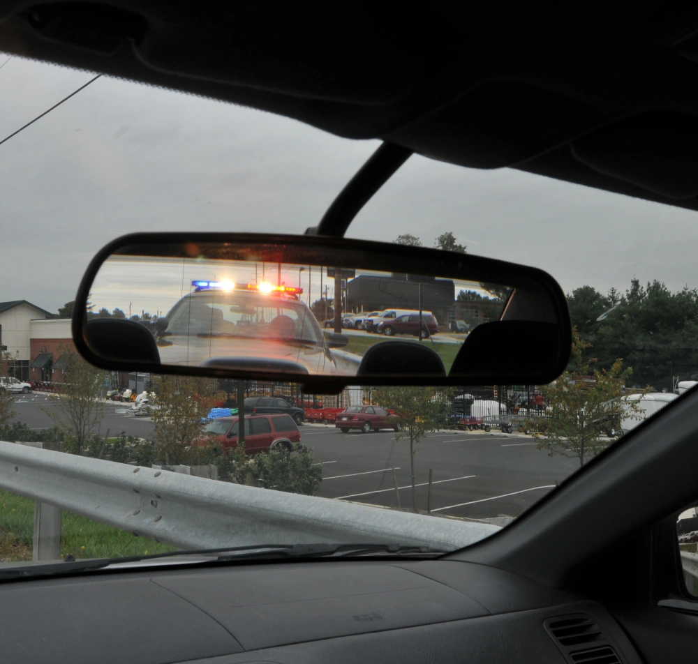 Police car with flashing lights visible in the rearview mirror, parked in a lot with trees and vehicles.