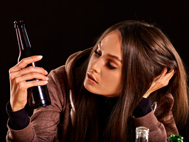 Woman in brown hoodie holding dark beer bottle against black background.