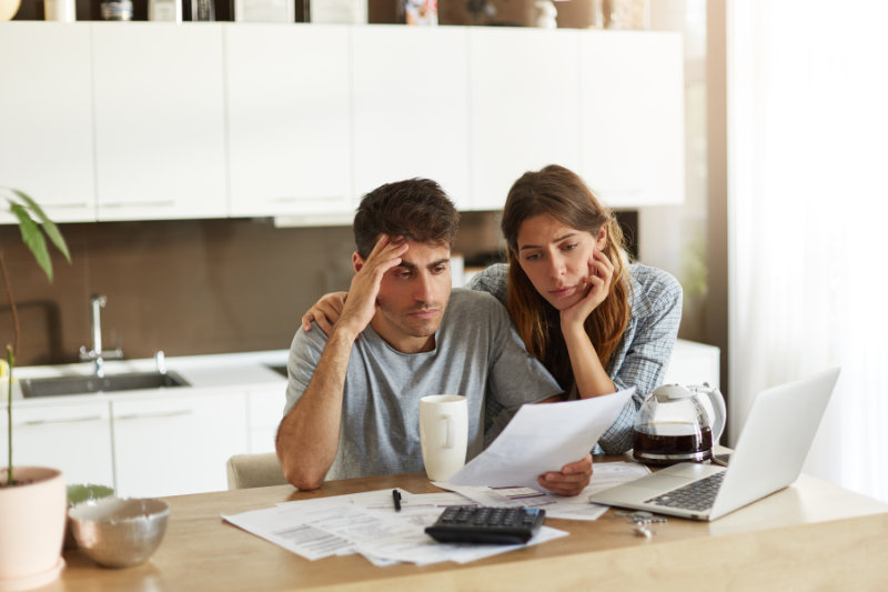A worried couple reviews bills and paperwork at a kitchen table, with a laptop and coffee nearby.