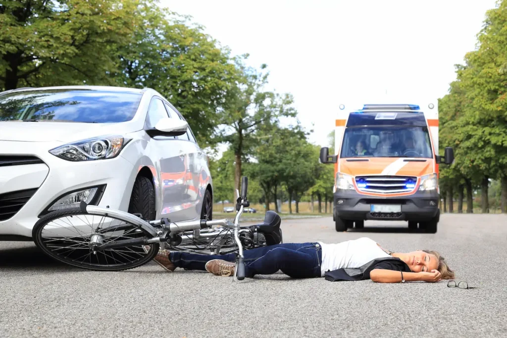 A woman lies on the road next to a fallen bicycle, with a white car nearby and an ambulance approaching.