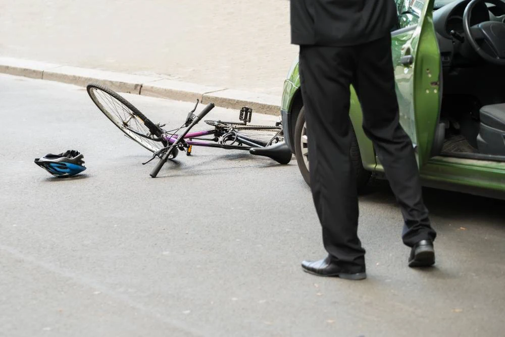 Bicycle lying on the road with a blue helmet nearby; a person in a suit stands next to a green car.