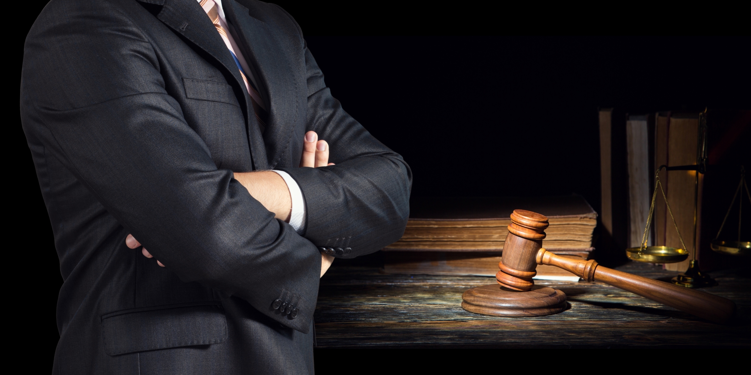 A man in a suit stands with arms crossed beside a wooden gavel and legal books on a dark background.