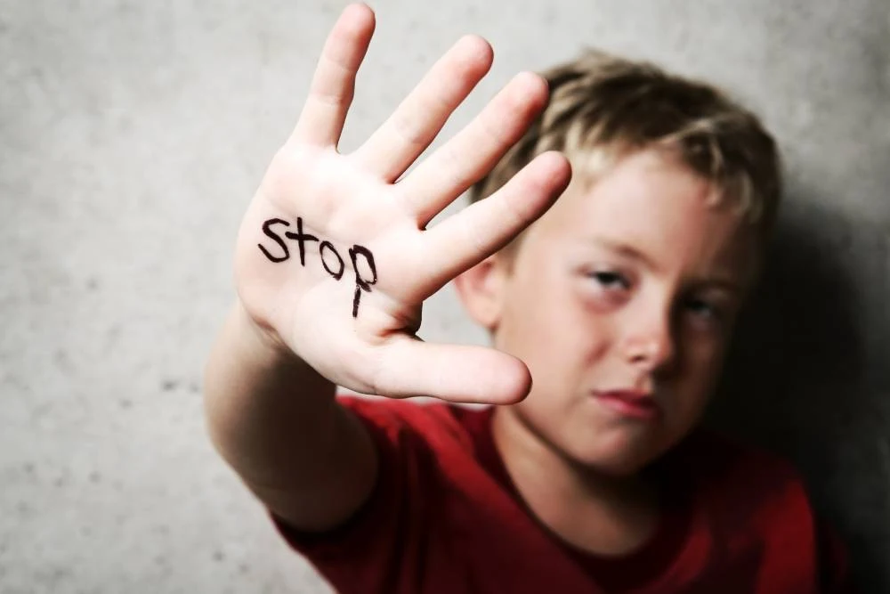 Child with a serious expression holds up a hand with "stop" written on it, against a gray background.