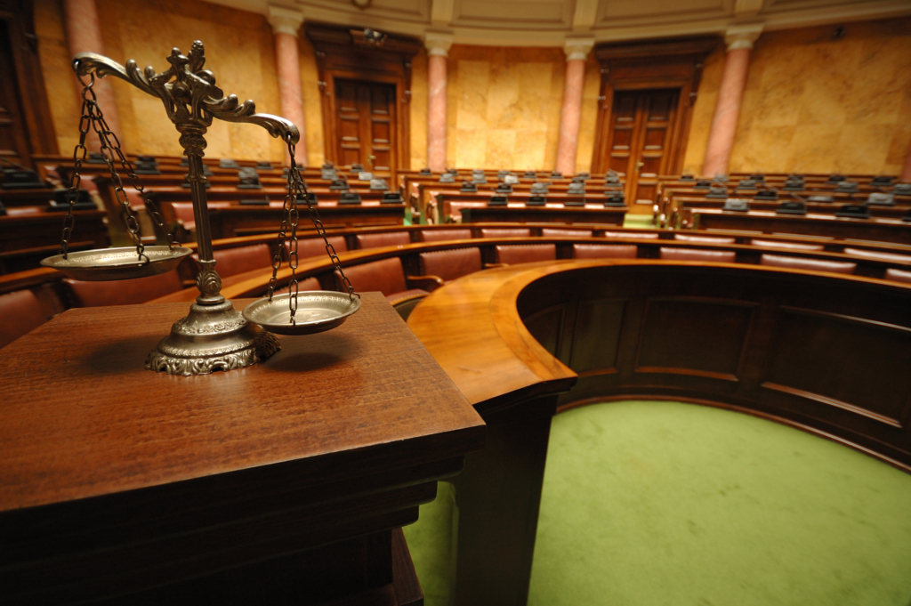Scales of justice on a wooden podium in an empty courtroom with brown leather seating and green carpet.