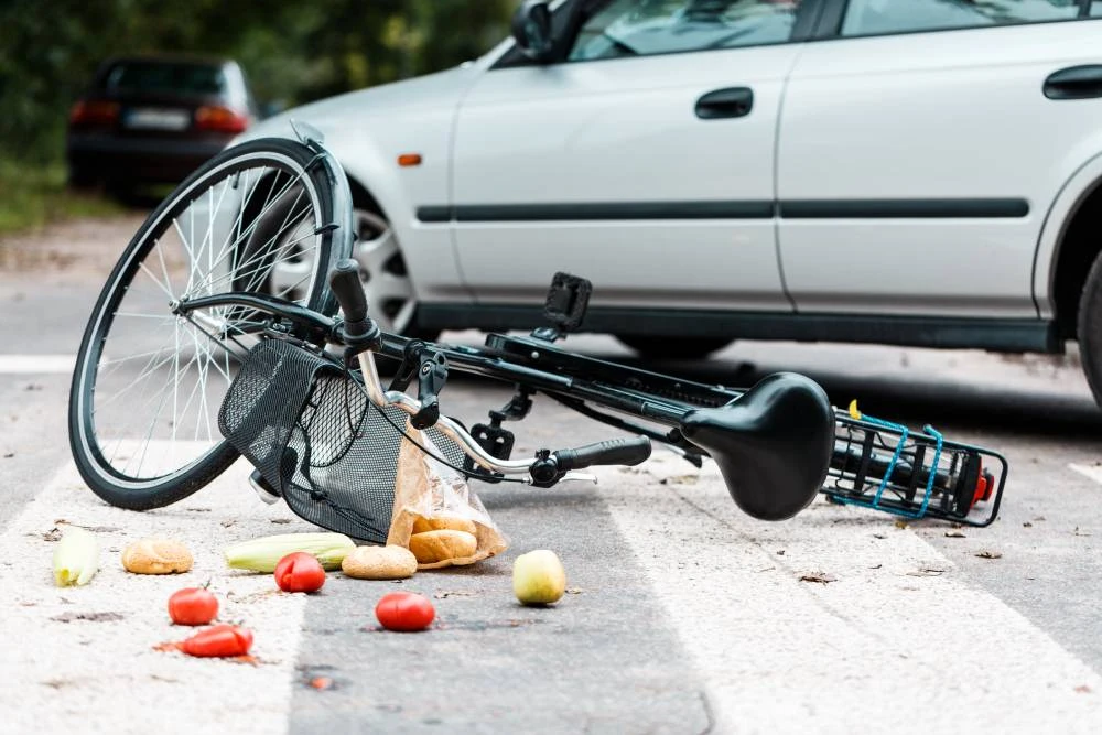 Bicycle on its side with scattered vegetables and bread on the road near a parked car.