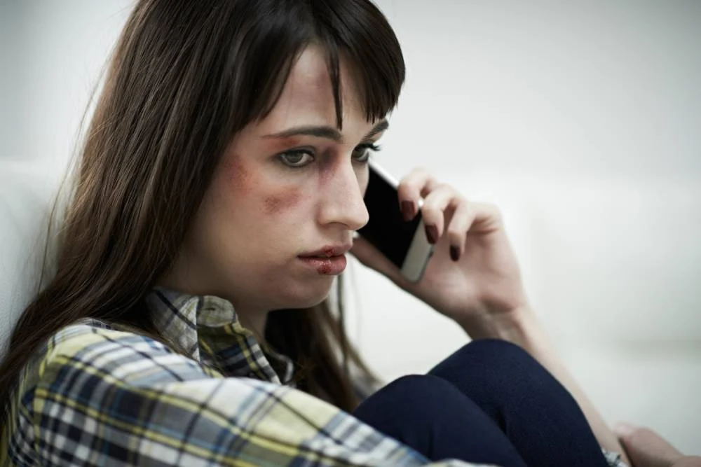 Woman with bruises and cuts, sitting on a couch, talking on a smartphone, wearing a plaid shirt.