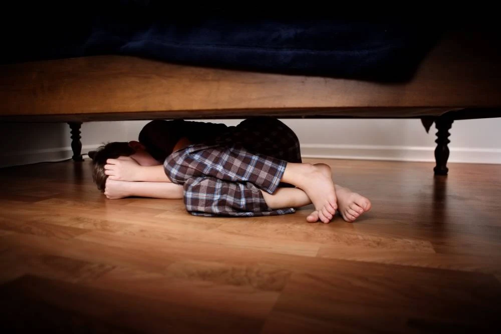 Child curled up under a bed, wearing plaid pajamas, with bare feet visible on wooden floor.