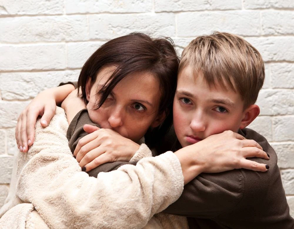 A woman and a boy embrace tightly, both looking distressed against a white brick wall.