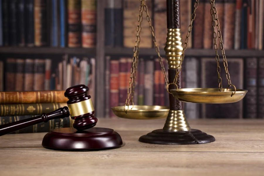 Brown gavel and scales of justice on a wooden table, with a blurred bookshelf in the background.