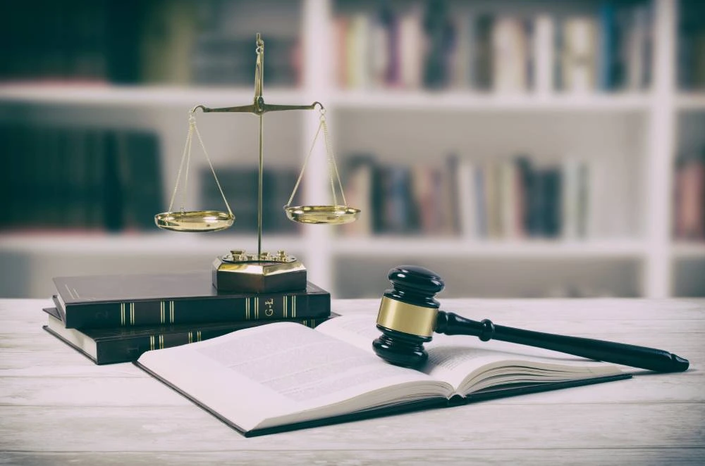 Brass scales of justice on books beside a black gavel and an open law book on a wooden table.