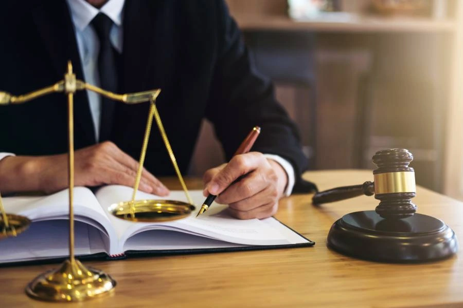 A person in a suit writes in a notebook, with a gavel and scales of justice on the table.