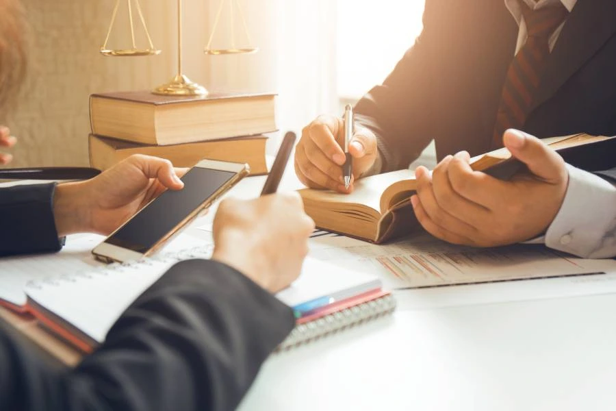 Two people in suits discussing legal documents, one holding a smartphone, with books and a scale in the background.