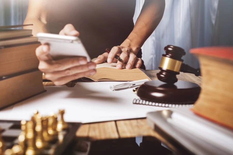 A person using a smartphone while writing in a book, surrounded by law books, a gavel, and a chess set.