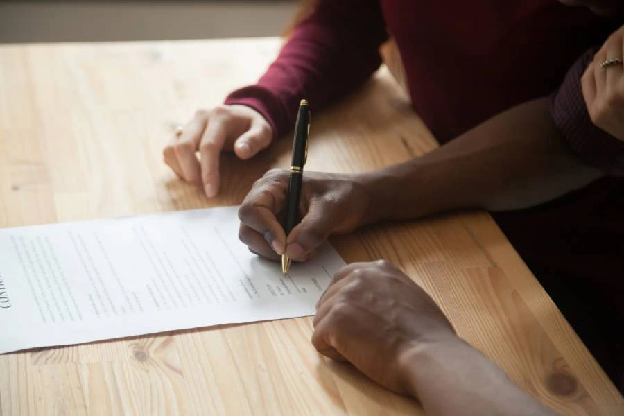 Hands signing a document on a wooden table, with a black pen and a partially visible contract.