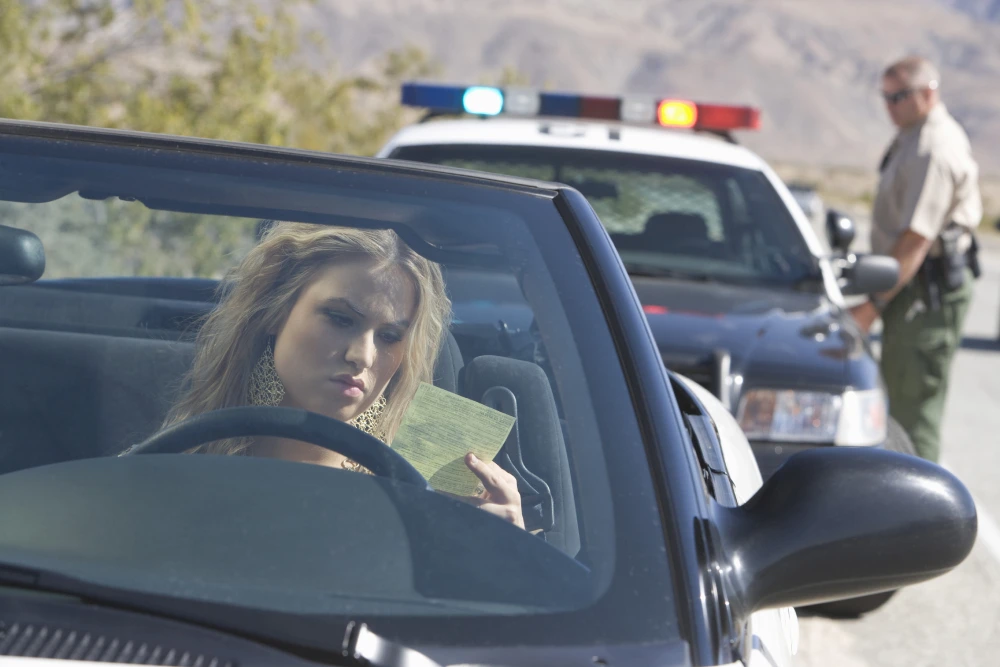Woman in a car holds a green ticket while a police officer approaches a patrol car in the background.