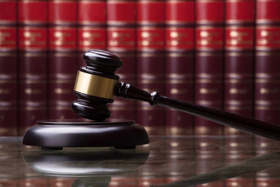 Wooden gavel with a gold band on a reflective surface, with red law books in the background.